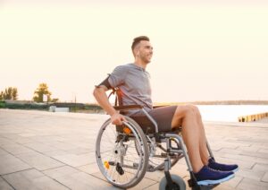 A white man in a wheelchair wearing a gray t-shirt and shorts with blue tennis shoes.